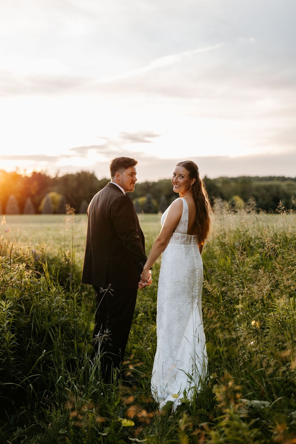 A bride and groom hold hands in a sunlit field at golden hour, bride smiling back in white lace gown.