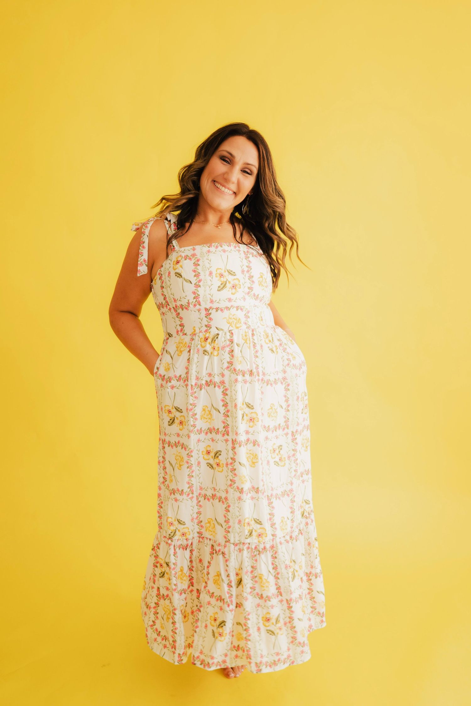 Woman in floral maxi dress with tie straps posing against yellow background, smiling.