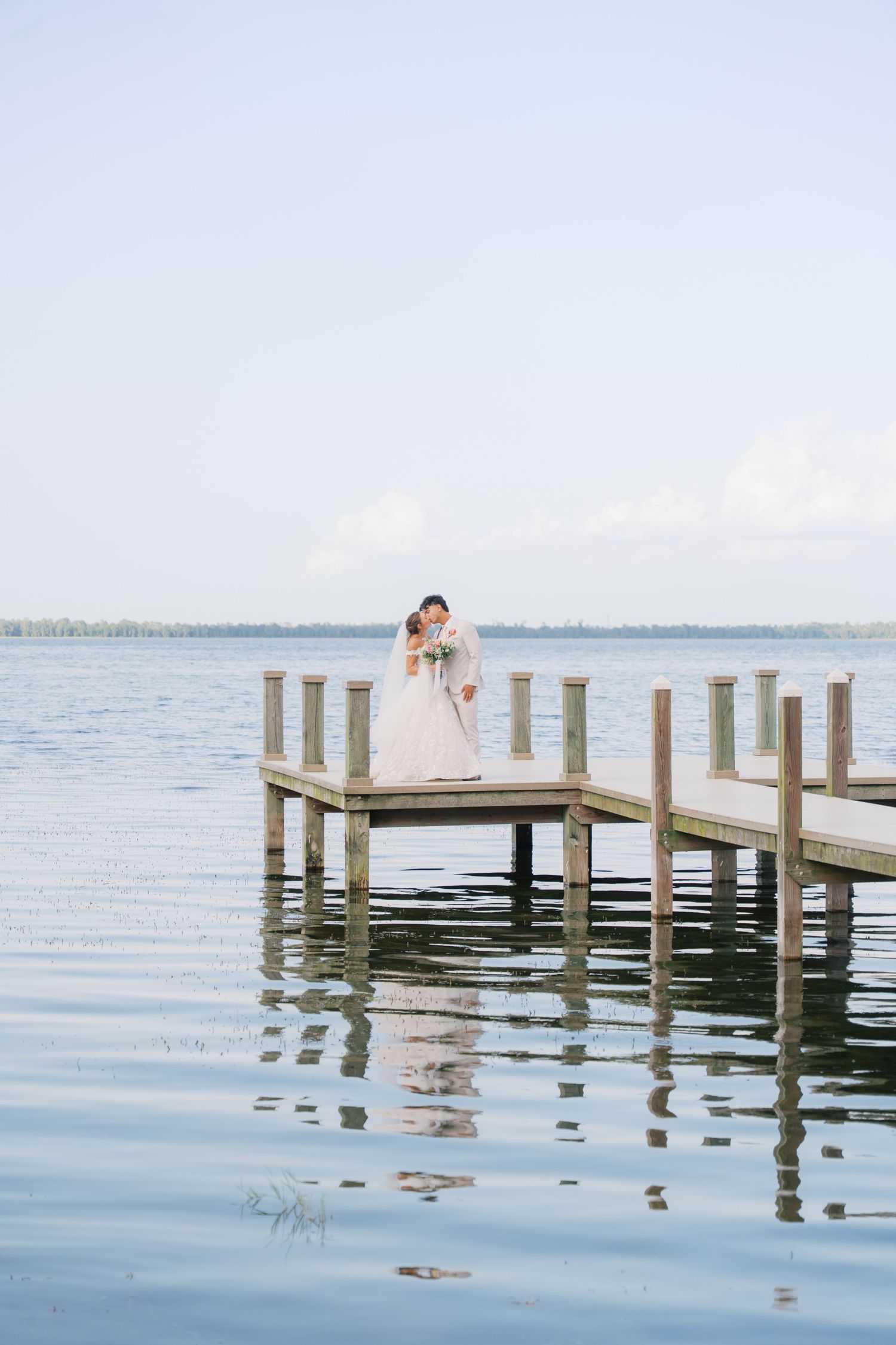 Bride and groom share a romantic dip kiss on a wooden dock at sunset overlooking a serene lake.
