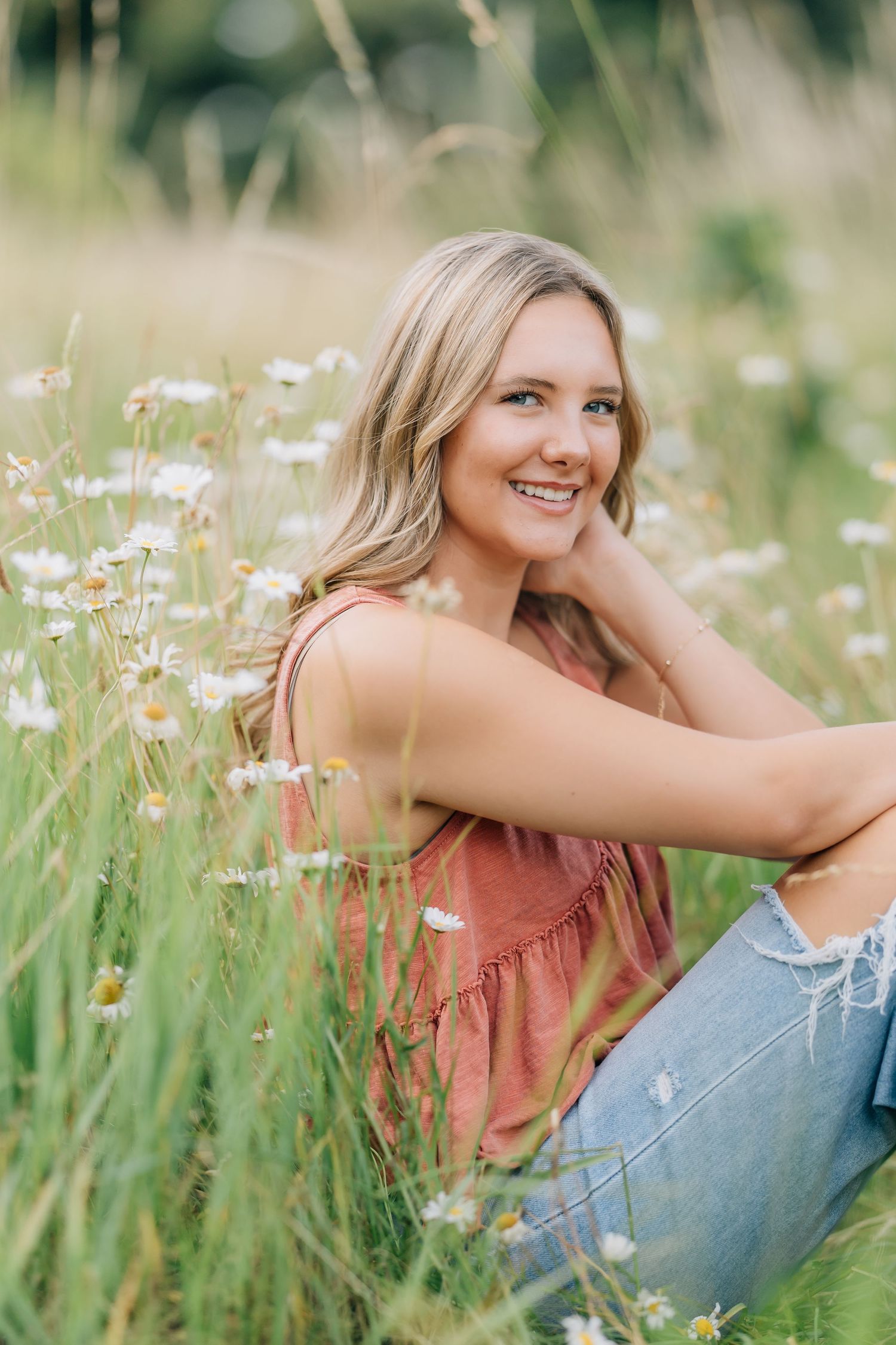Young blonde woman smiling in a wildflower meadow, wearing a pink top and ripped jeans, sitting among white daisies.