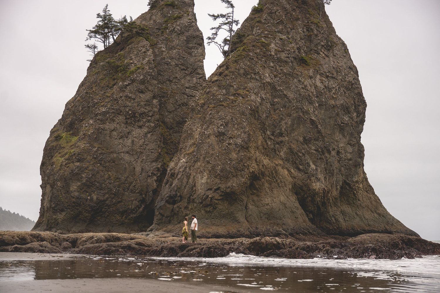 A couple stands in front of split rock on Rialto beach in Olympic National Park.