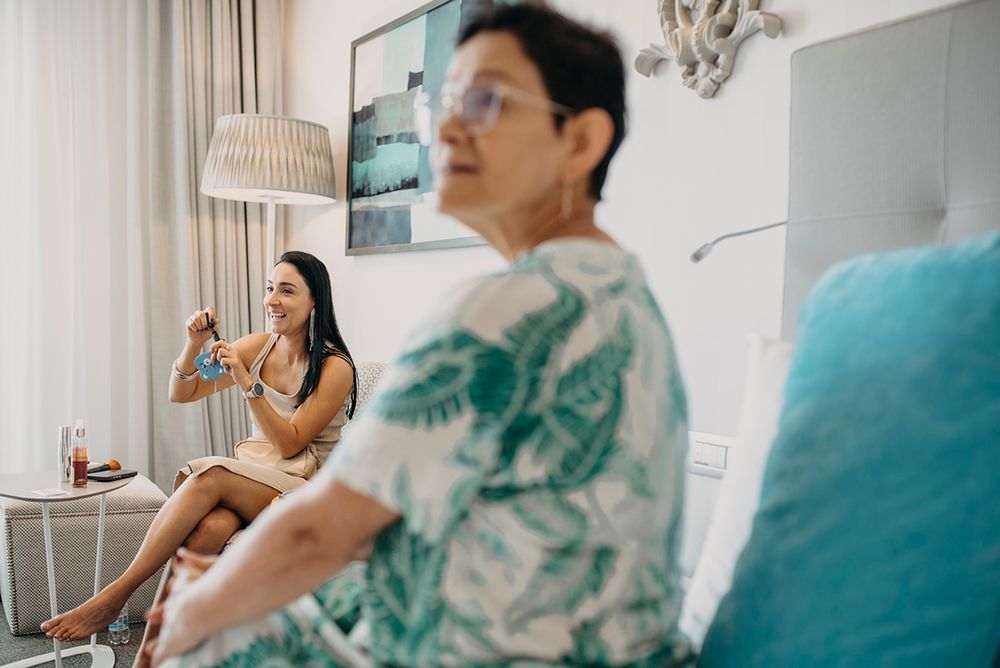Two women relaxing in a bright hotel room, one sitting and holding a drink while the other rests in the foreground.