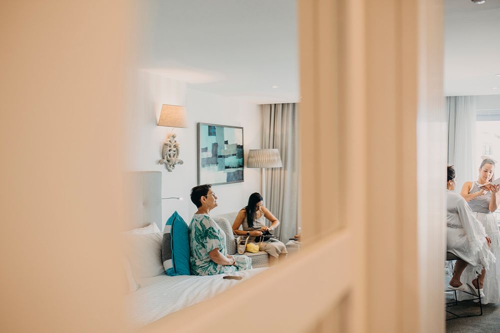 Two women relaxing in a bright hotel room, sitting on a bed while others chat in the background.