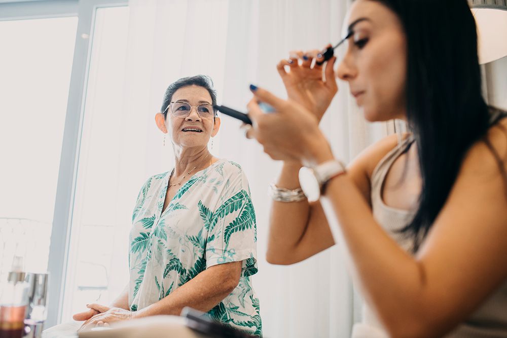 An older woman smiles warmly watching a young brunette woman apply mascara during bridal wedding preparations.