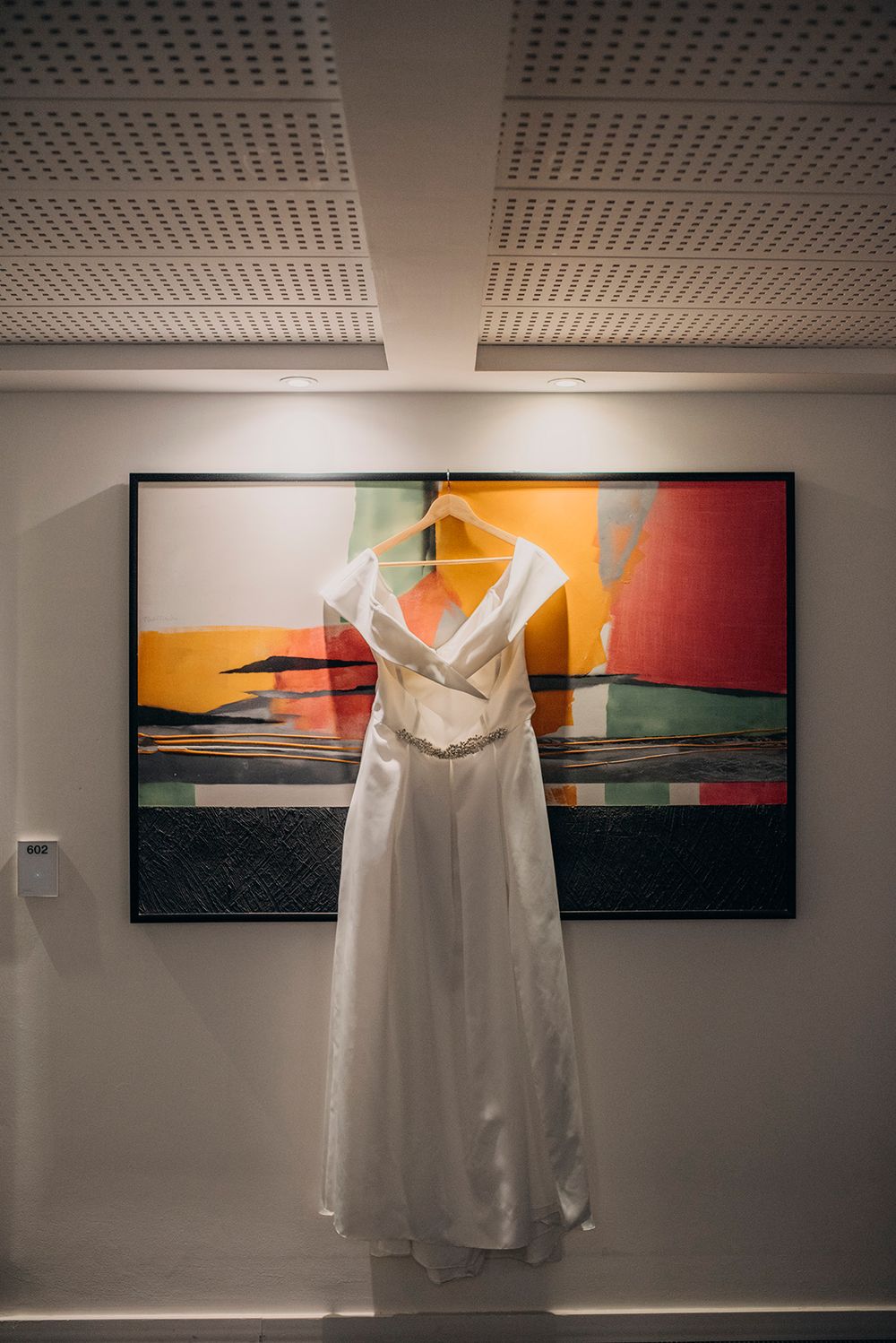 White wedding dress hanging on a wooden hanger against a colorful abstract painting in a modern gallery.