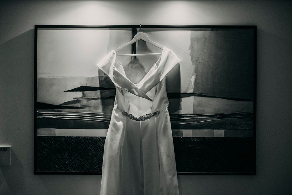 Elegant white wedding dress hanging on a hanger in front of a framed artwork in black and white.