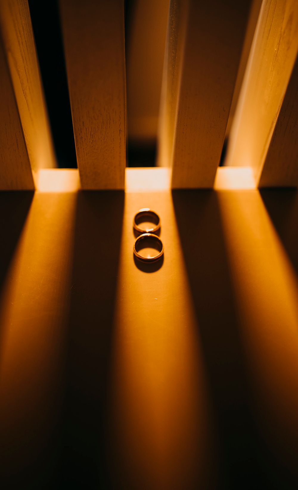 Two gold wedding rings glowing in warm light between wooden slats, creating a romantic shadow pattern.