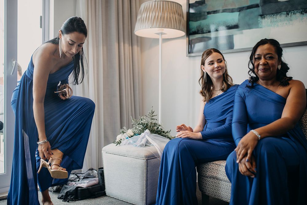 Three bridesmaids in blue dresses prepare for a wedding, one adjusting her shoes while two others sit smiling.