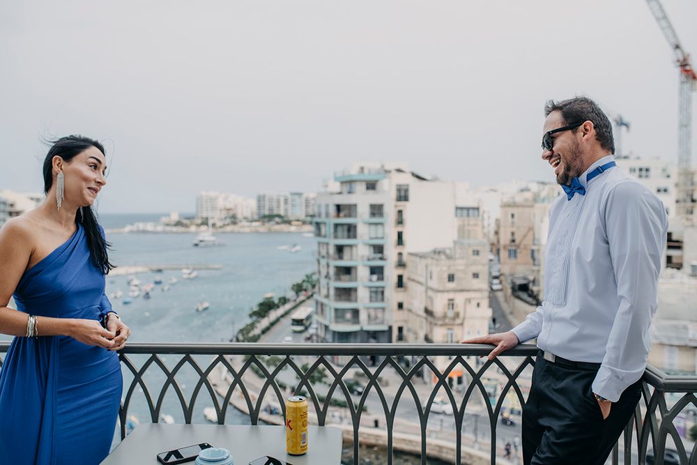 A woman in a blue dress and a man in sunglasses chat on a rooftop balcony overlooking a coastal city.