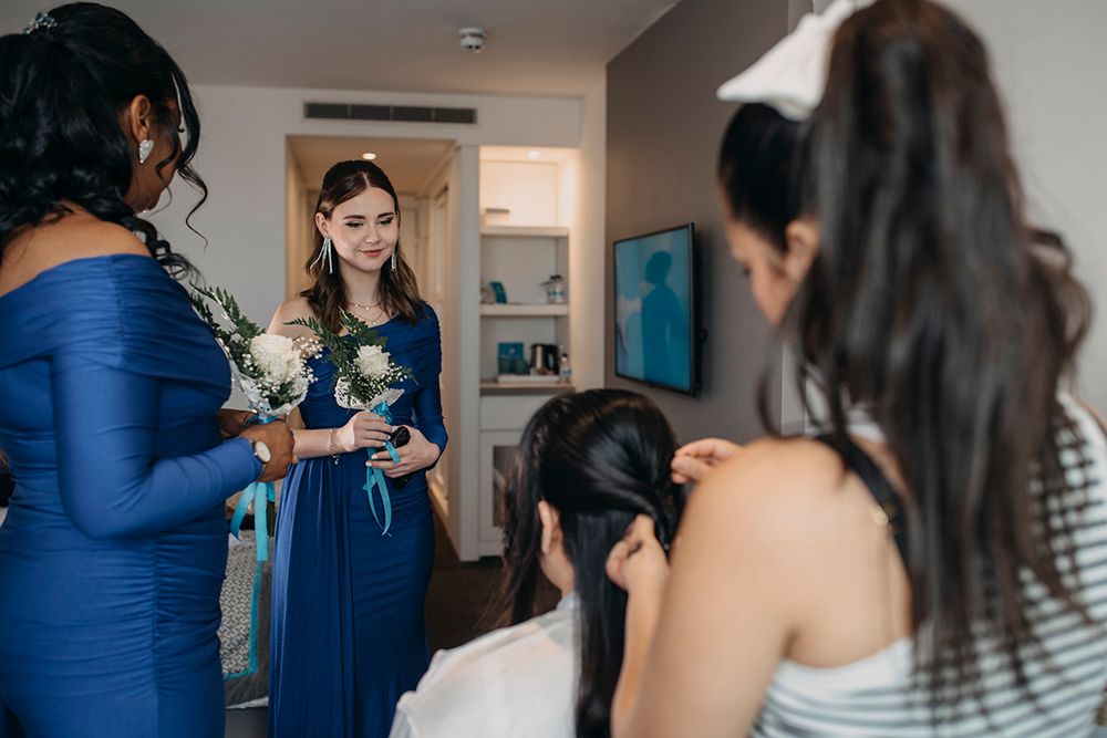 Bridesmaids in blue dresses gather in a hotel room, holding bouquets while a stylist arranges hair before the wedding.