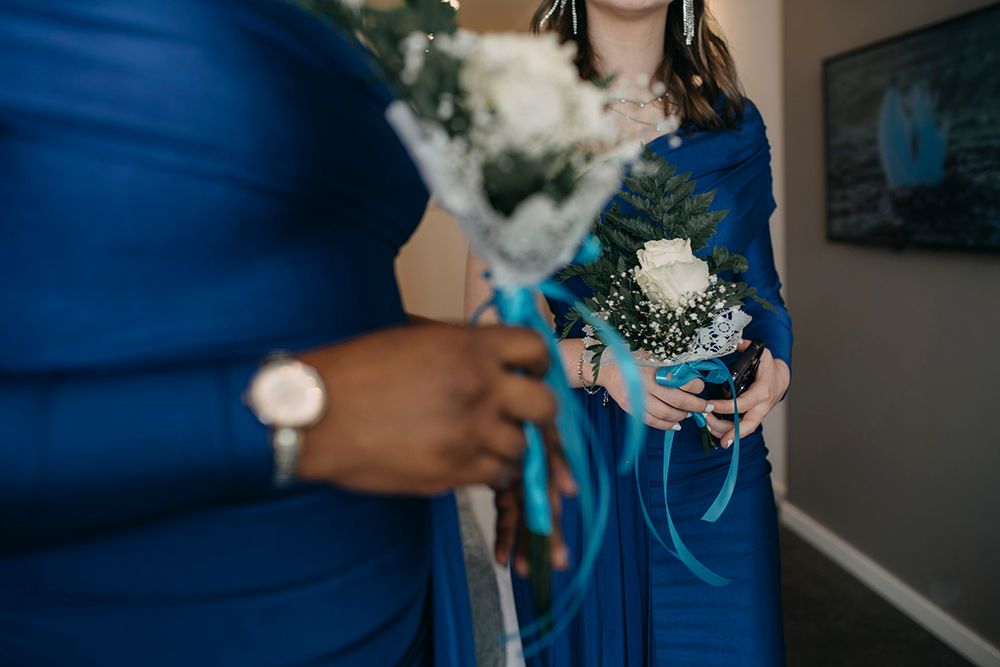 Bridesmaids in royal blue dresses holding white floral bouquets with teal ribbons at a wedding ceremony.