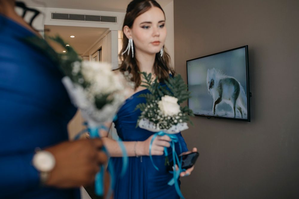 Bridesmaid in blue dress holding white bouquet stands near framed artwork at wedding venue.