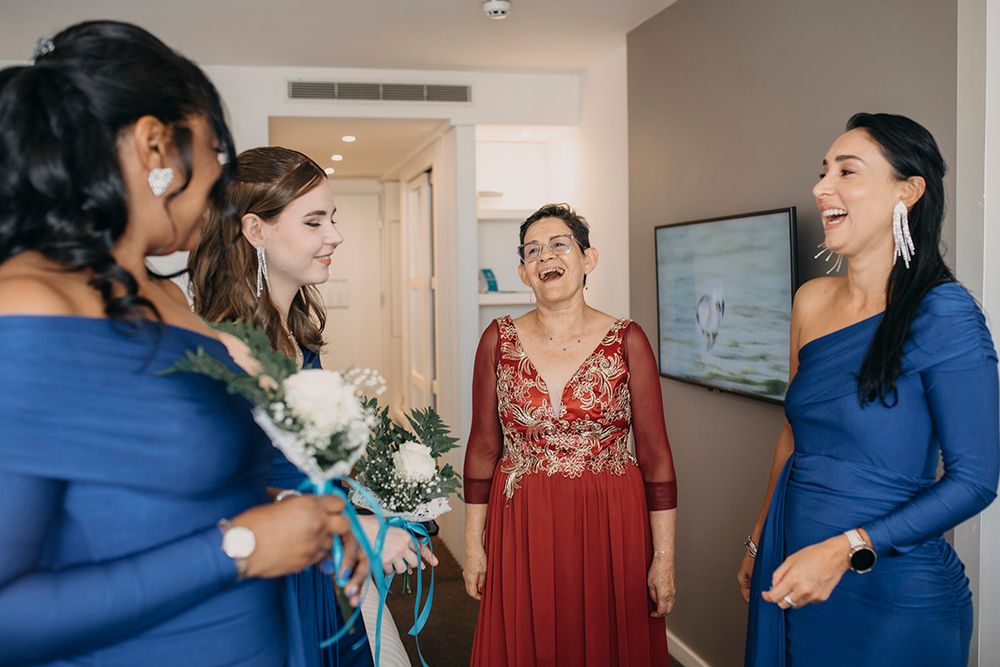 Bridesmaids in blue dresses laugh with woman in red gown holding white bouquet in bright hotel room.