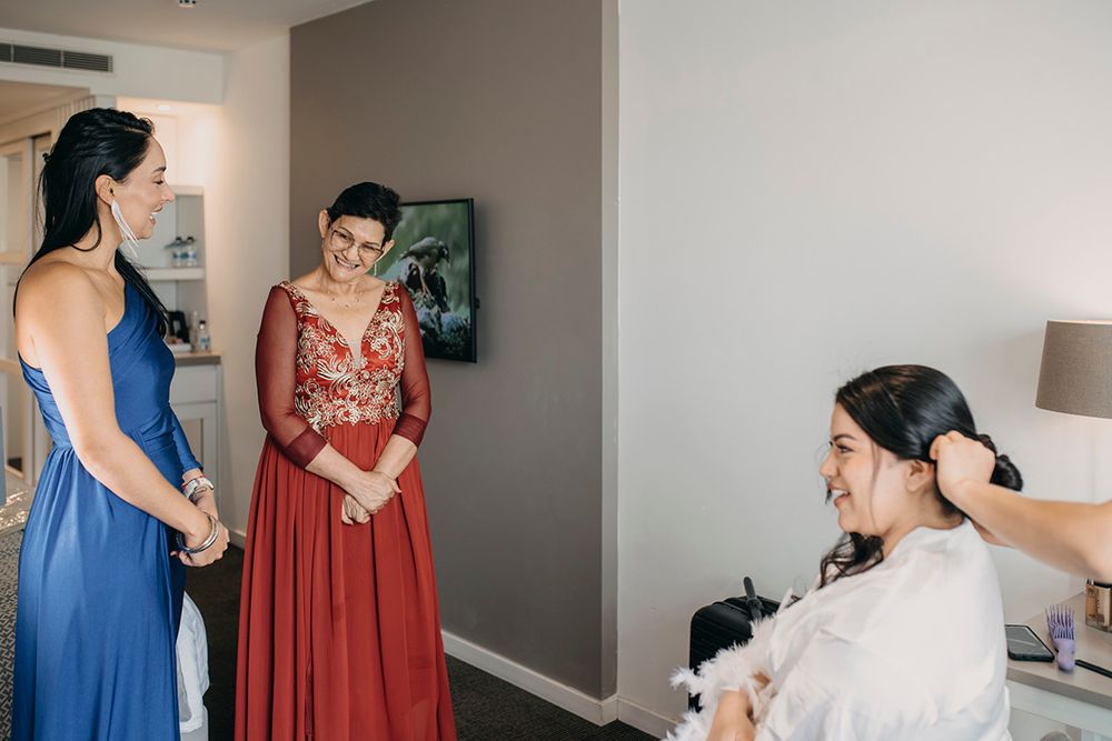 Bride smiling during hair styling while bridesmaids in blue and red dresses look on in hotel room.