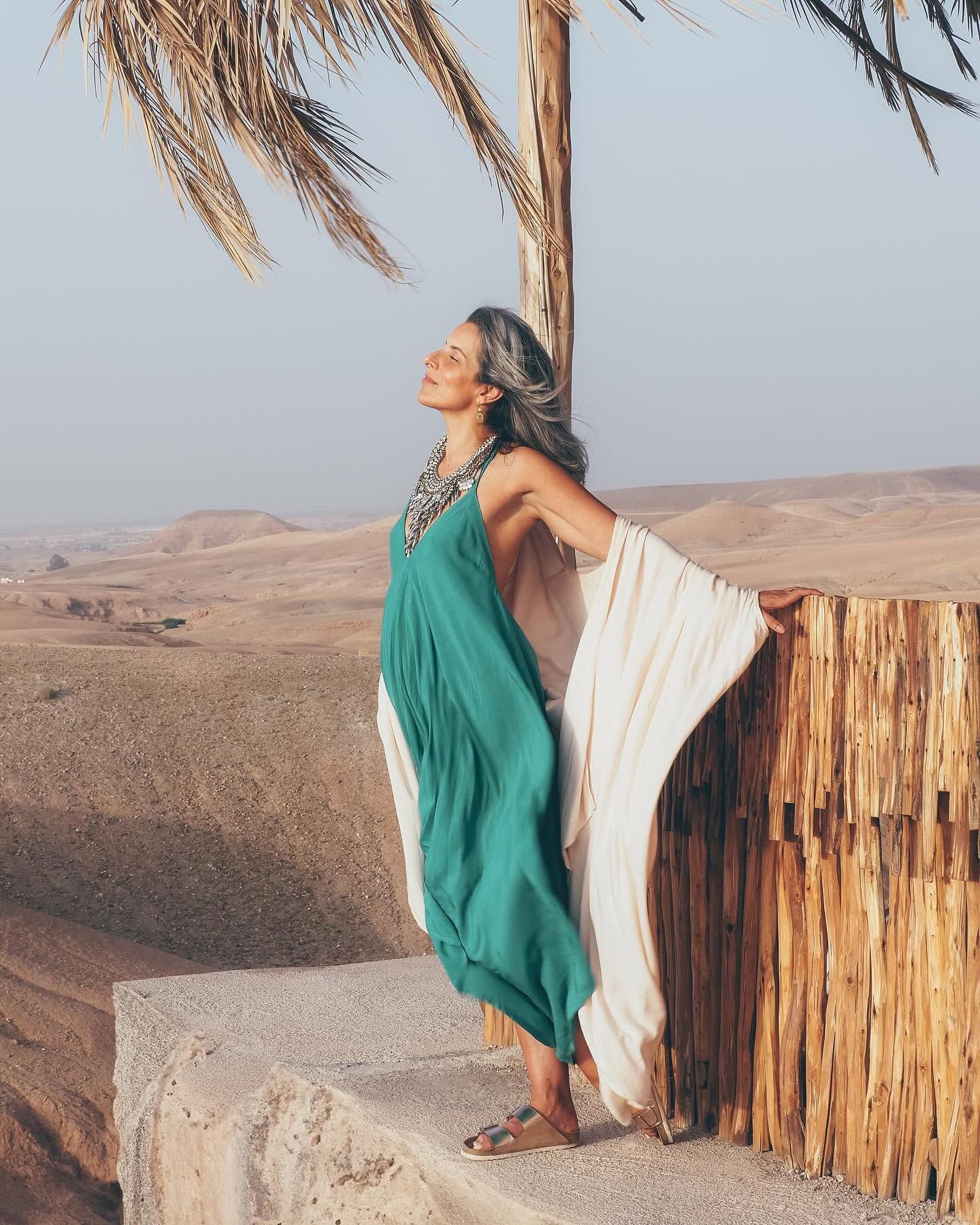 Woman in teal kaftan dress poses by palm tree overlooking vast desert landscape at golden hour.