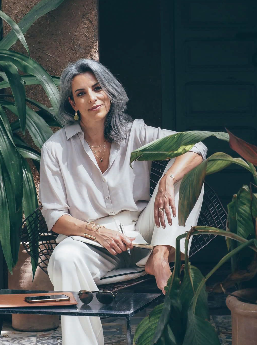 Elegant woman with gray hair in white outfit sitting among lush green tropical plants in a stylish outdoor setting.