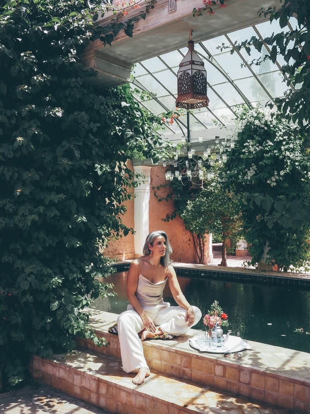 Woman in white outfit sitting poolside in a lush green Moroccan riad with ornate lantern overhead.