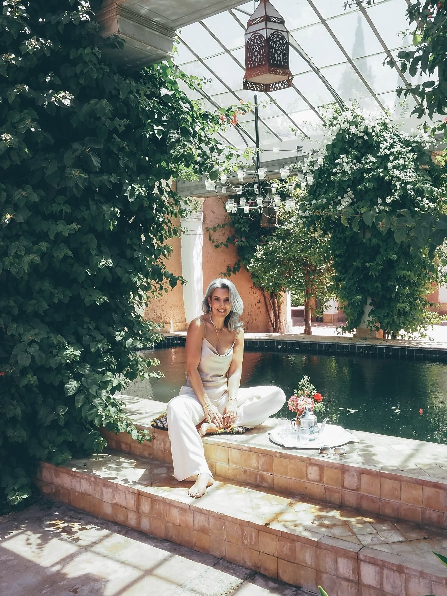 Woman in white outfit sitting by an indoor pool surrounded by lush greenery and Moroccan decor.