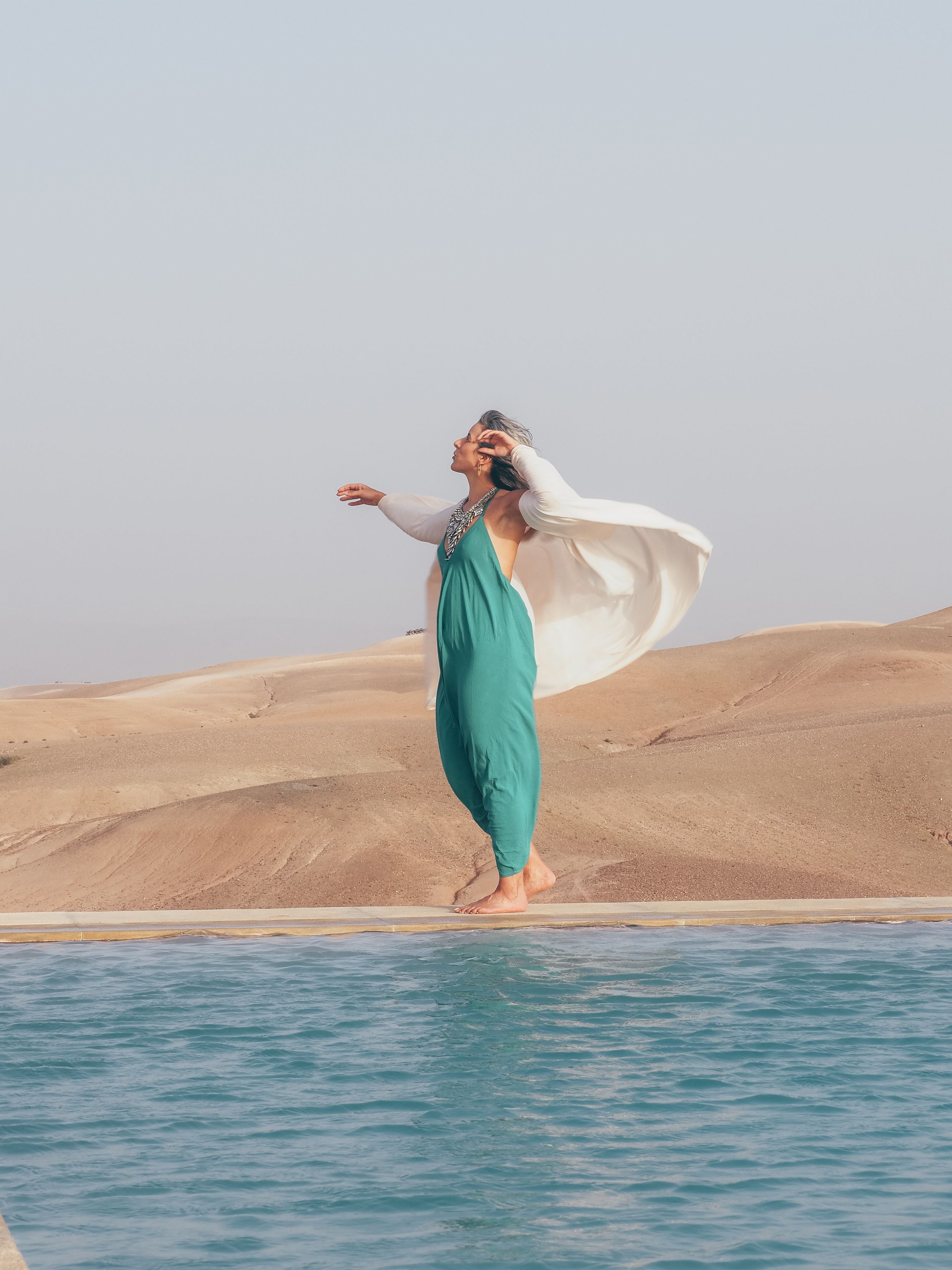 Woman in teal jumpsuit and white flowing scarf stands by infinity pool edge with sand dunes in desert background.