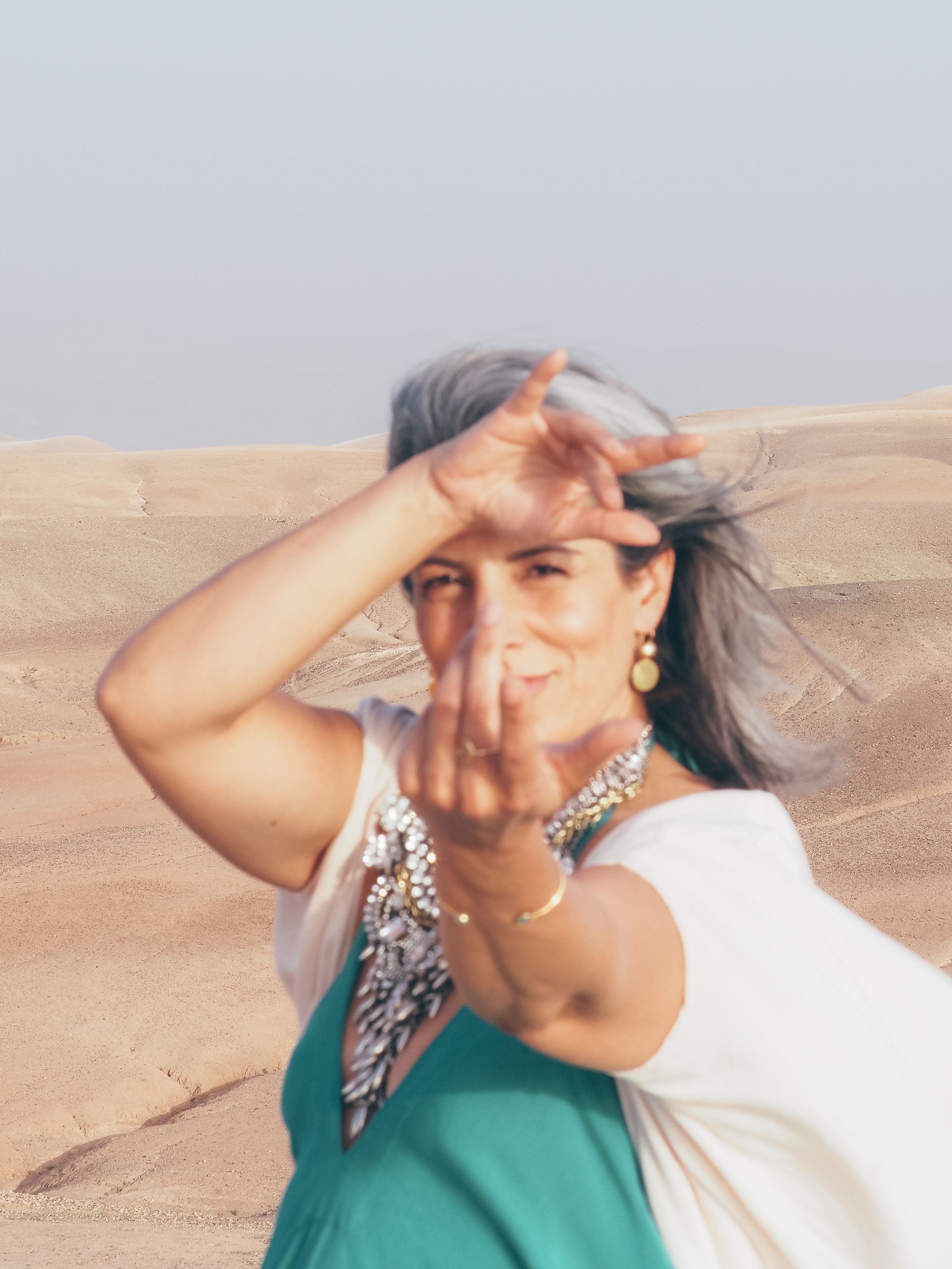 Woman in teal dress and white shawl shielding eyes from sun in desert landscape, wearing gold jewelry.