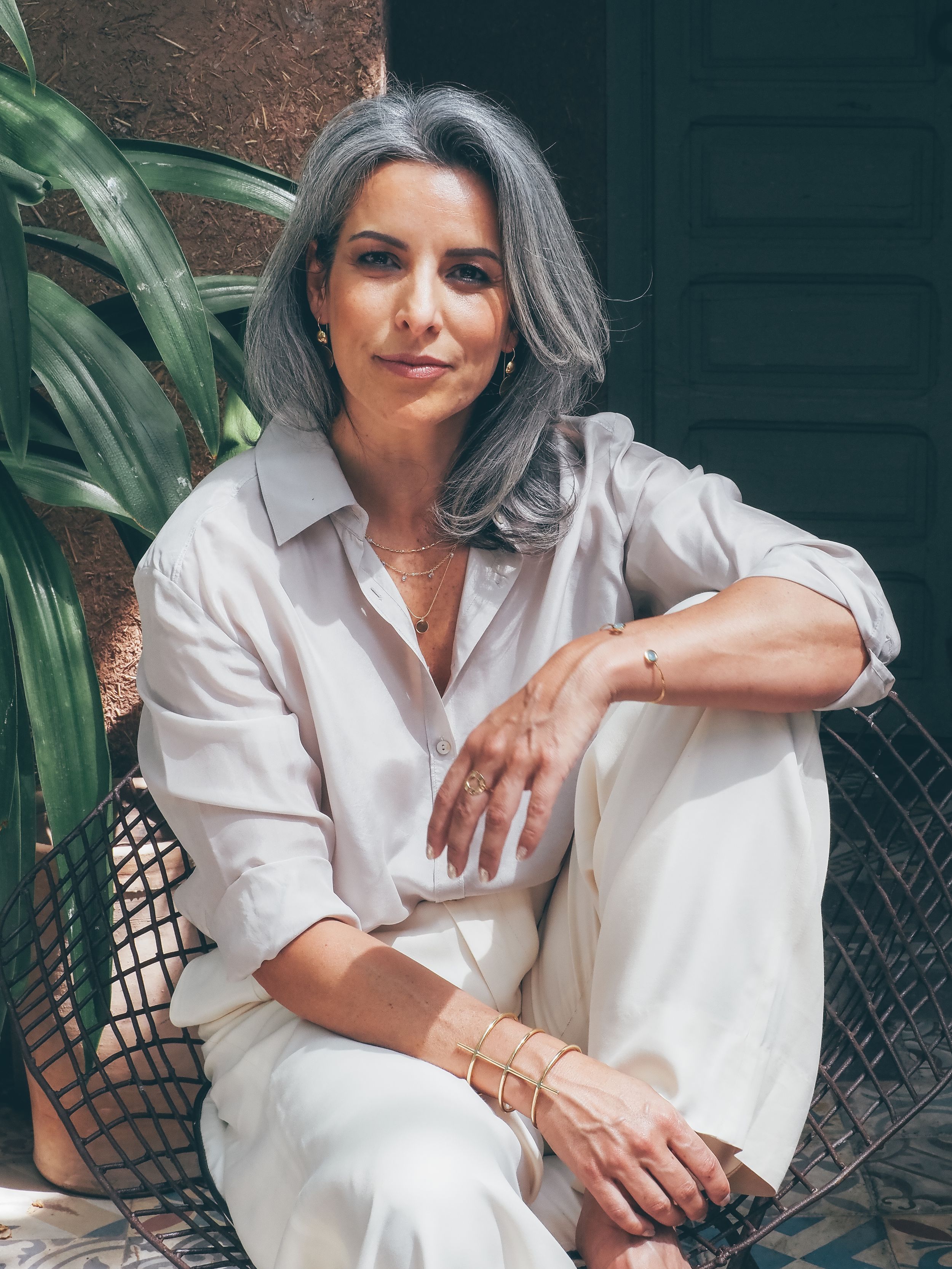 A stylish middle-aged woman with gray hair wearing a white linen outfit and gold bracelets sits relaxed near tropical plants.