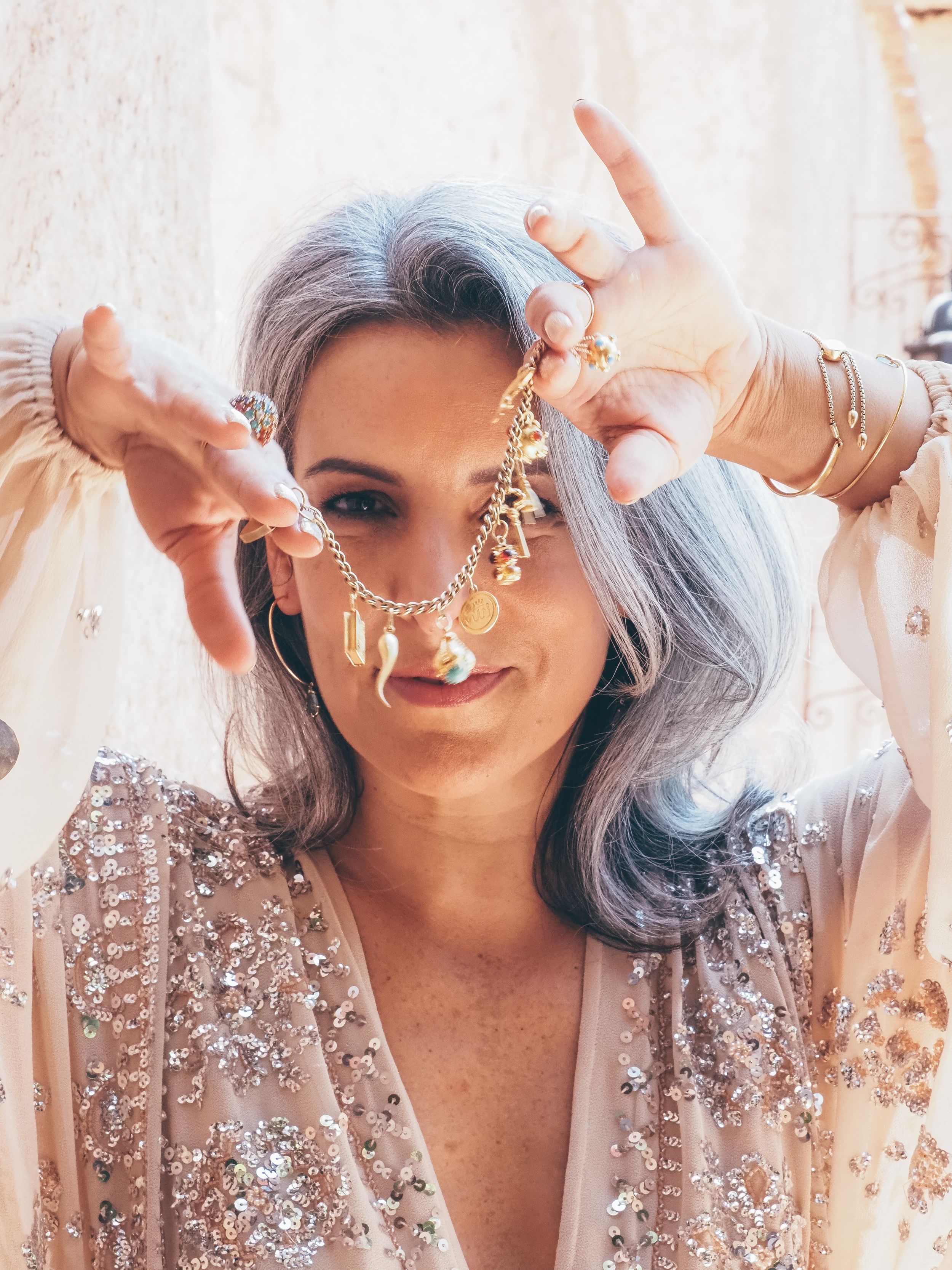 Woman with silver hair wearing gold jewelry and sequined dress, holding a gold chain necklace near her face.