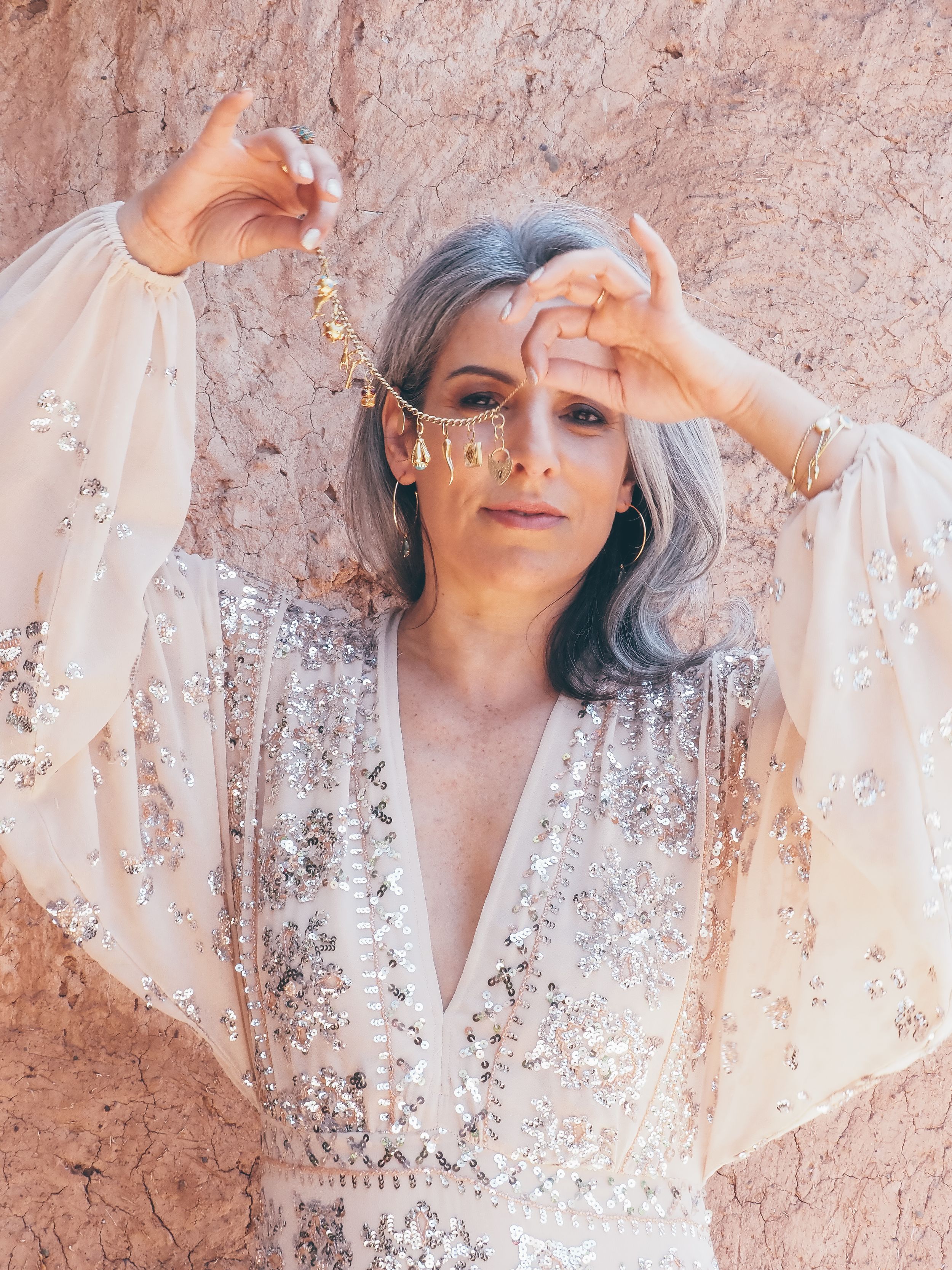Elegant woman with silver hair wearing a sequined gown and gold head chain jewelry, posing against a sandy wall.