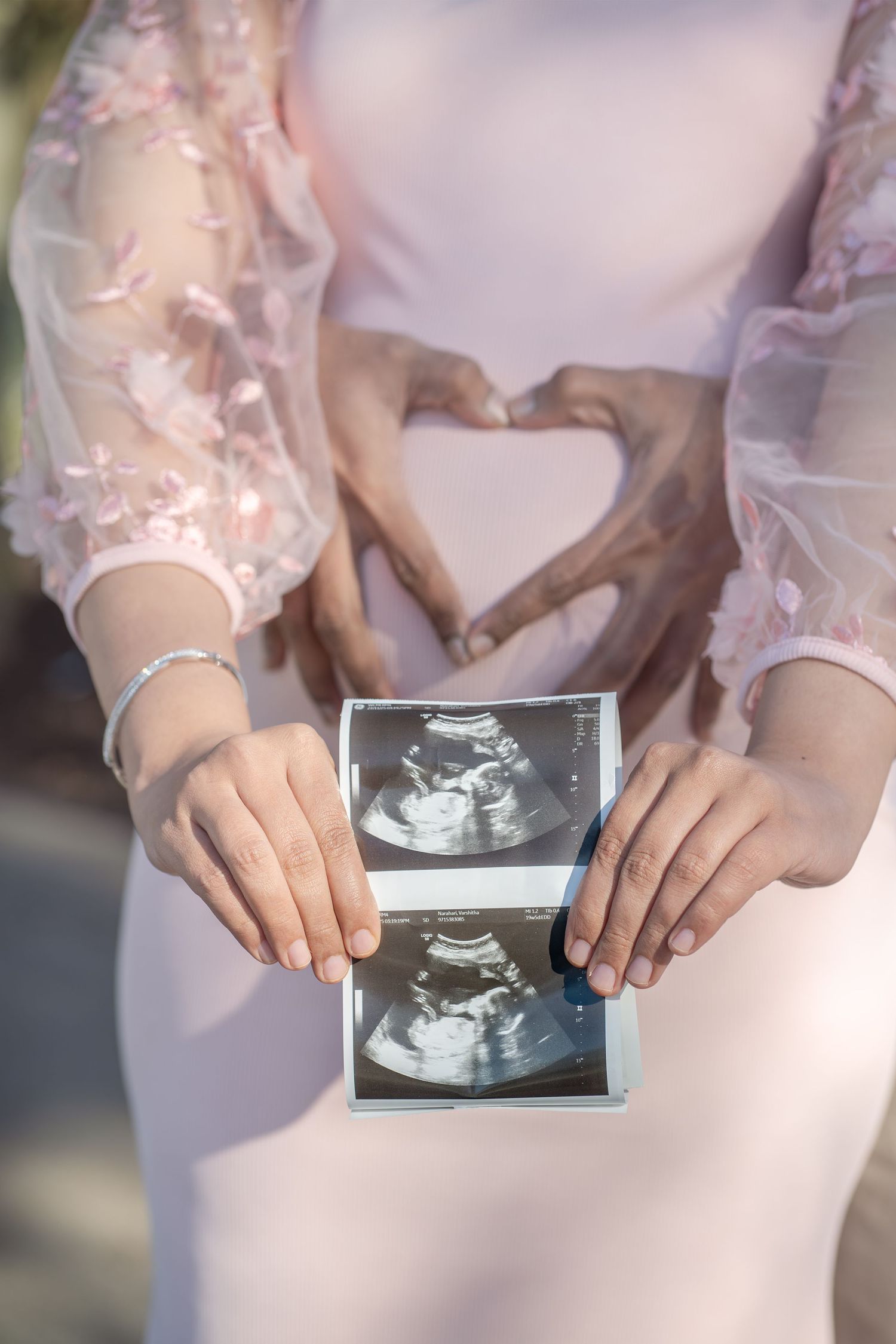 Pregnant woman in pink dress holding ultrasound scans while couple forms heart shape with hands over baby bump.