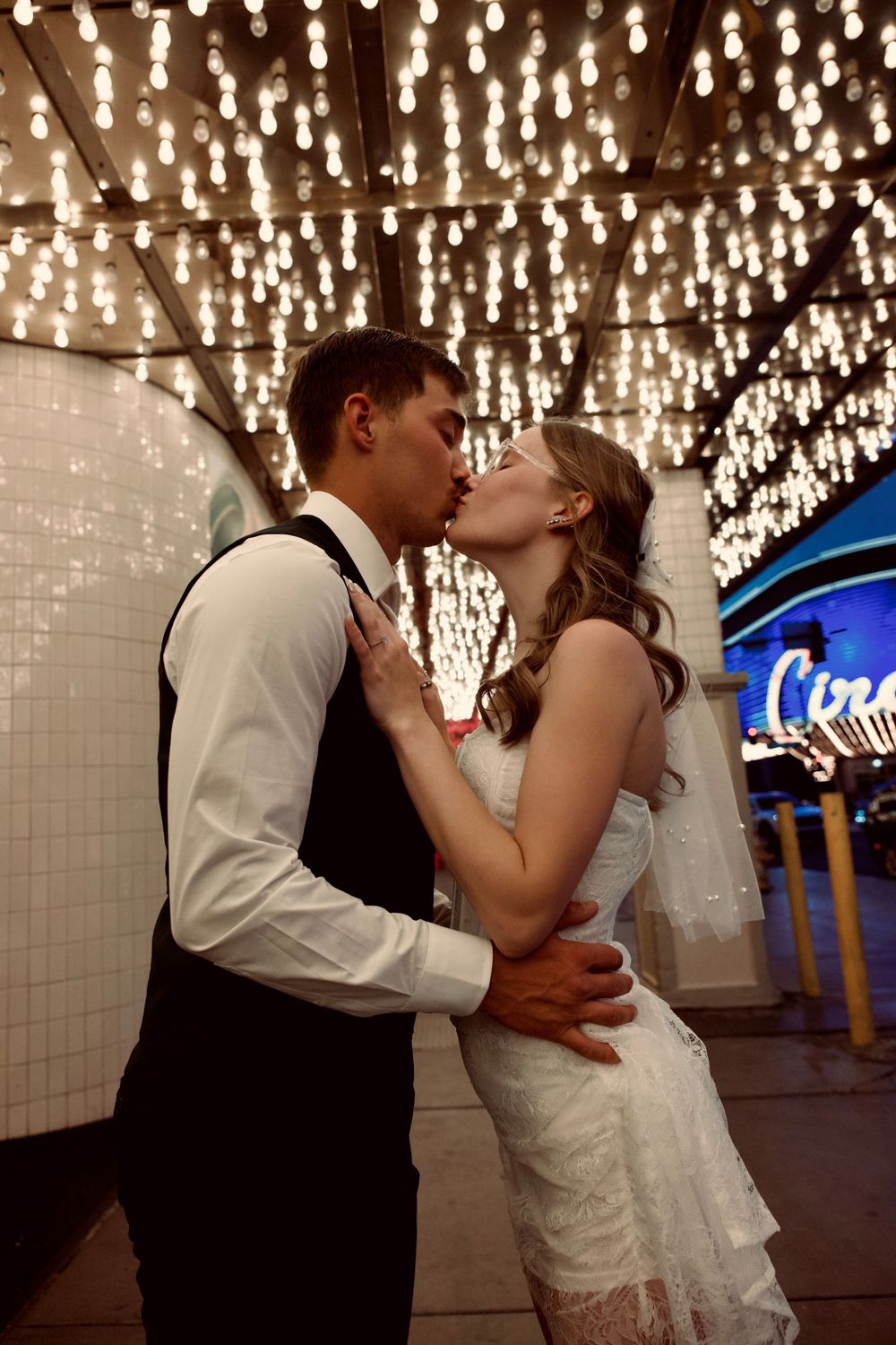 Bride and groom kissing passionately under sparkling string lights near a Las Vegas casino sign.