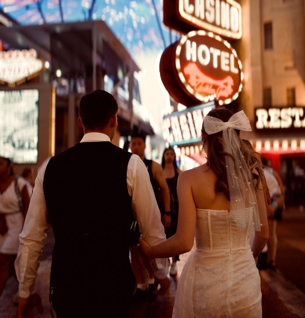 Bride and groom walking hand-in-hand past neon casino hotel signs on the Las Vegas Strip at night.