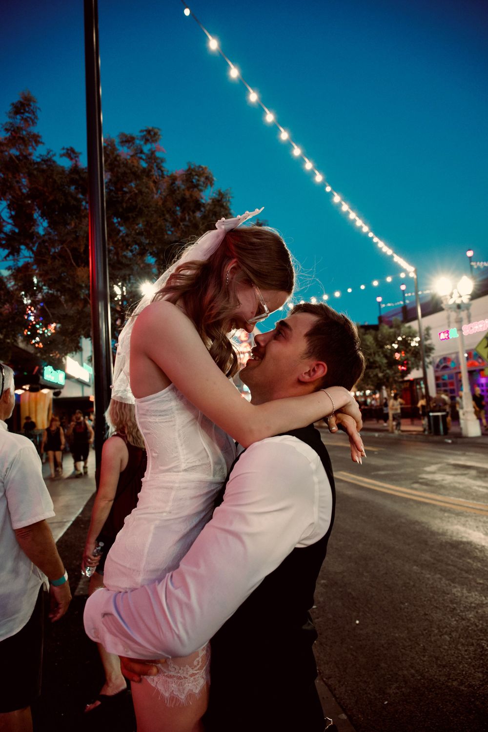 Groom lifting bride in wedding attire on a festive street at dusk with string lights overhead.