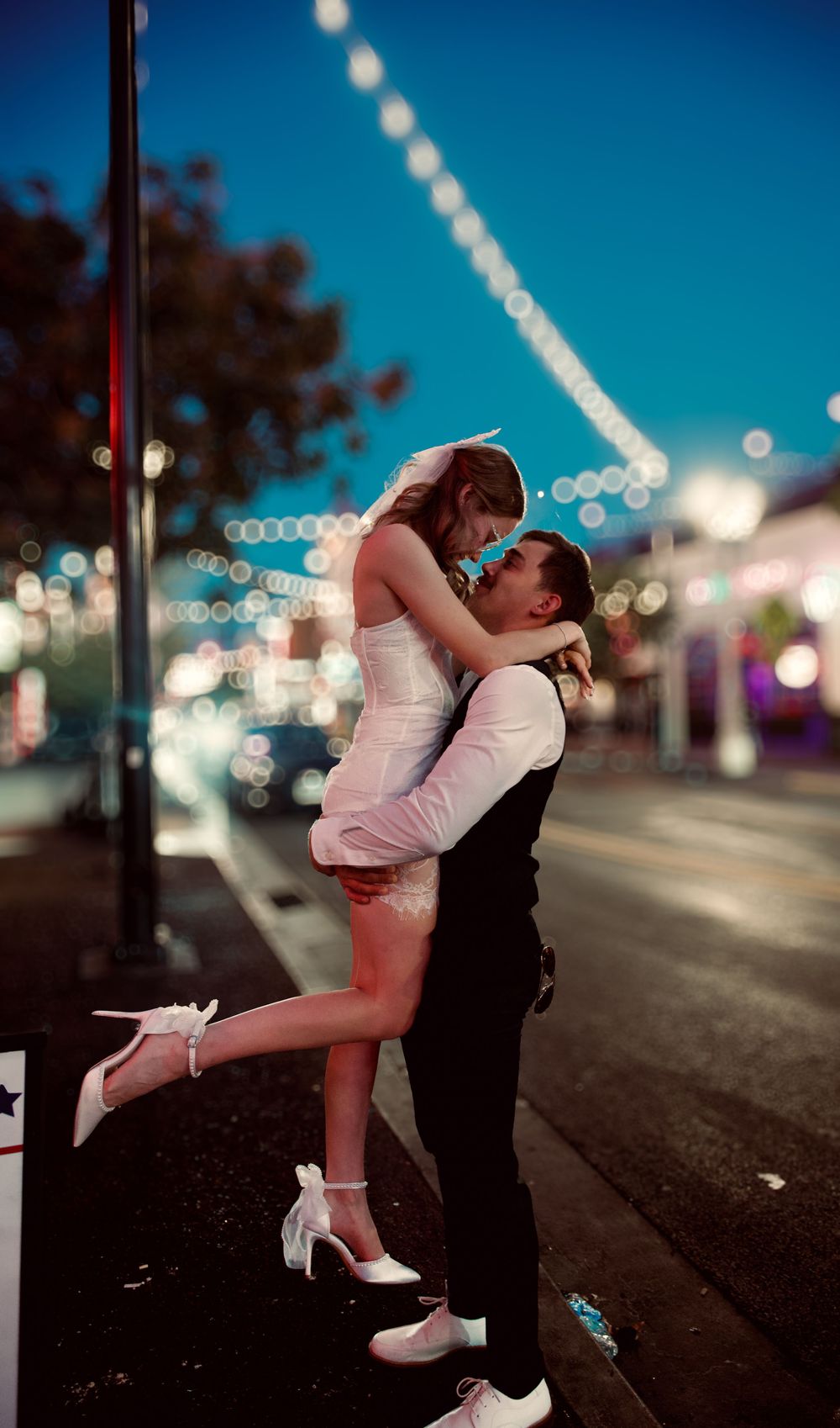 Groom lifting bride in white dress on city street at night with bokeh lights behind them.