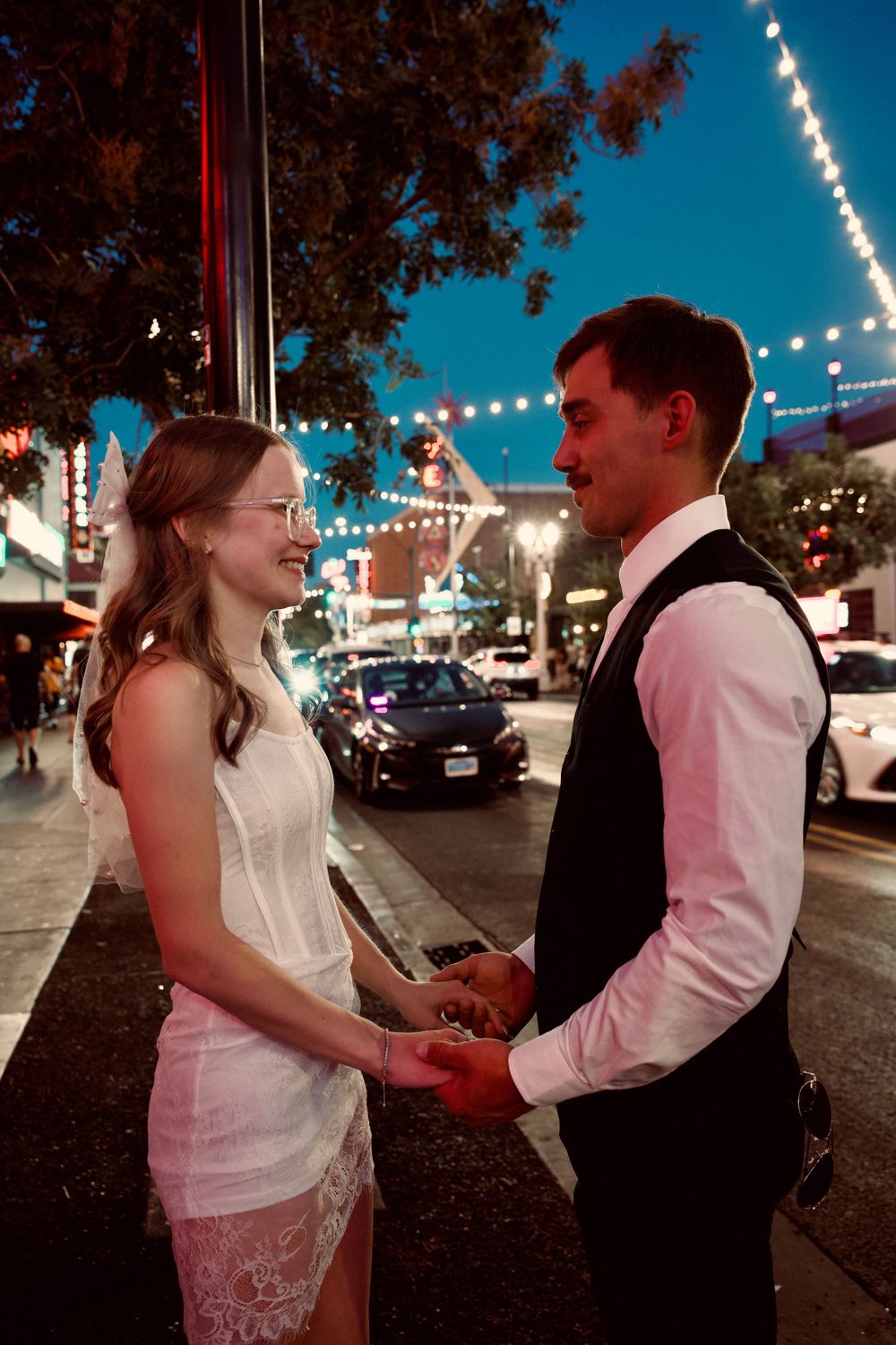 Bride and groom holding hands smiling on Las Vegas street at dusk with string lights.