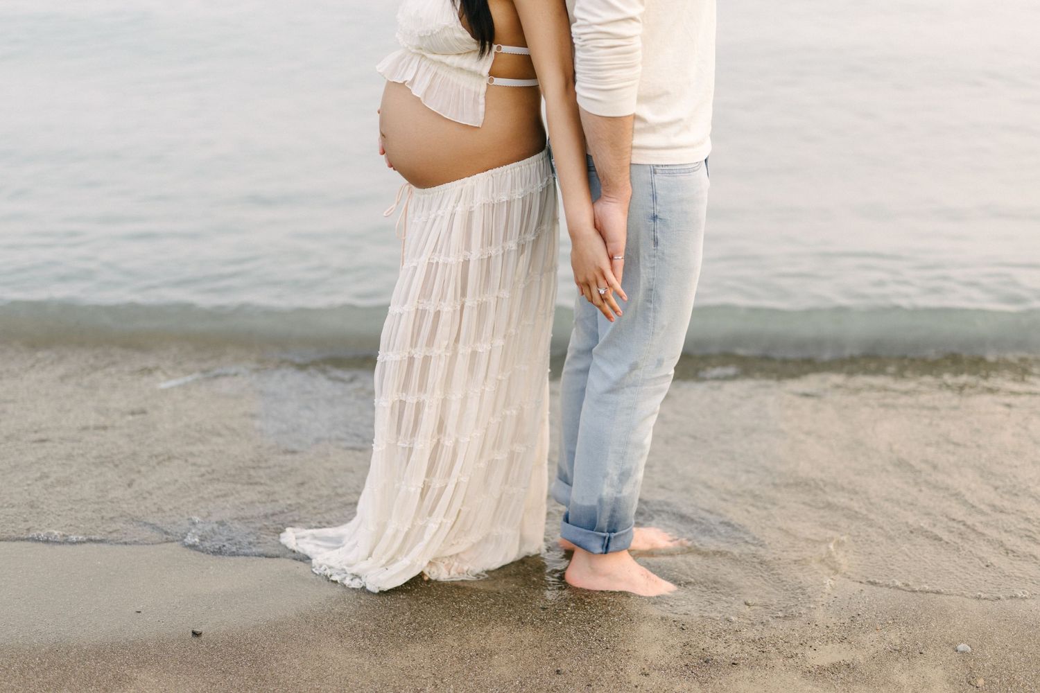 Expectant mother in a lace dress standing barefoot at the lake shore with her partner.