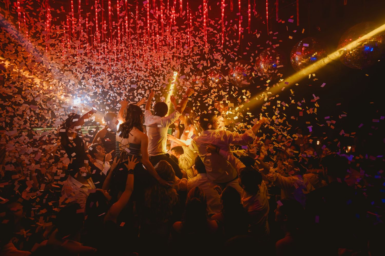 Bride lifted by guests amid golden confetti explosion under red lights at wedding reception.