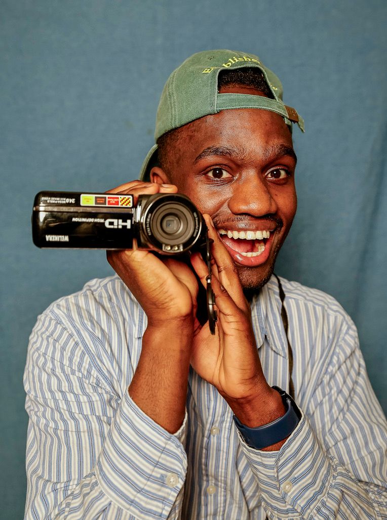 Smiling young man holding an HD video camera beside his face, wearing a backwards green cap and striped shirt.