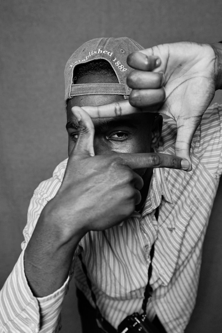 Black and white portrait of a man framing his eye with his fingers, wearing a striped shirt and baseball cap.
