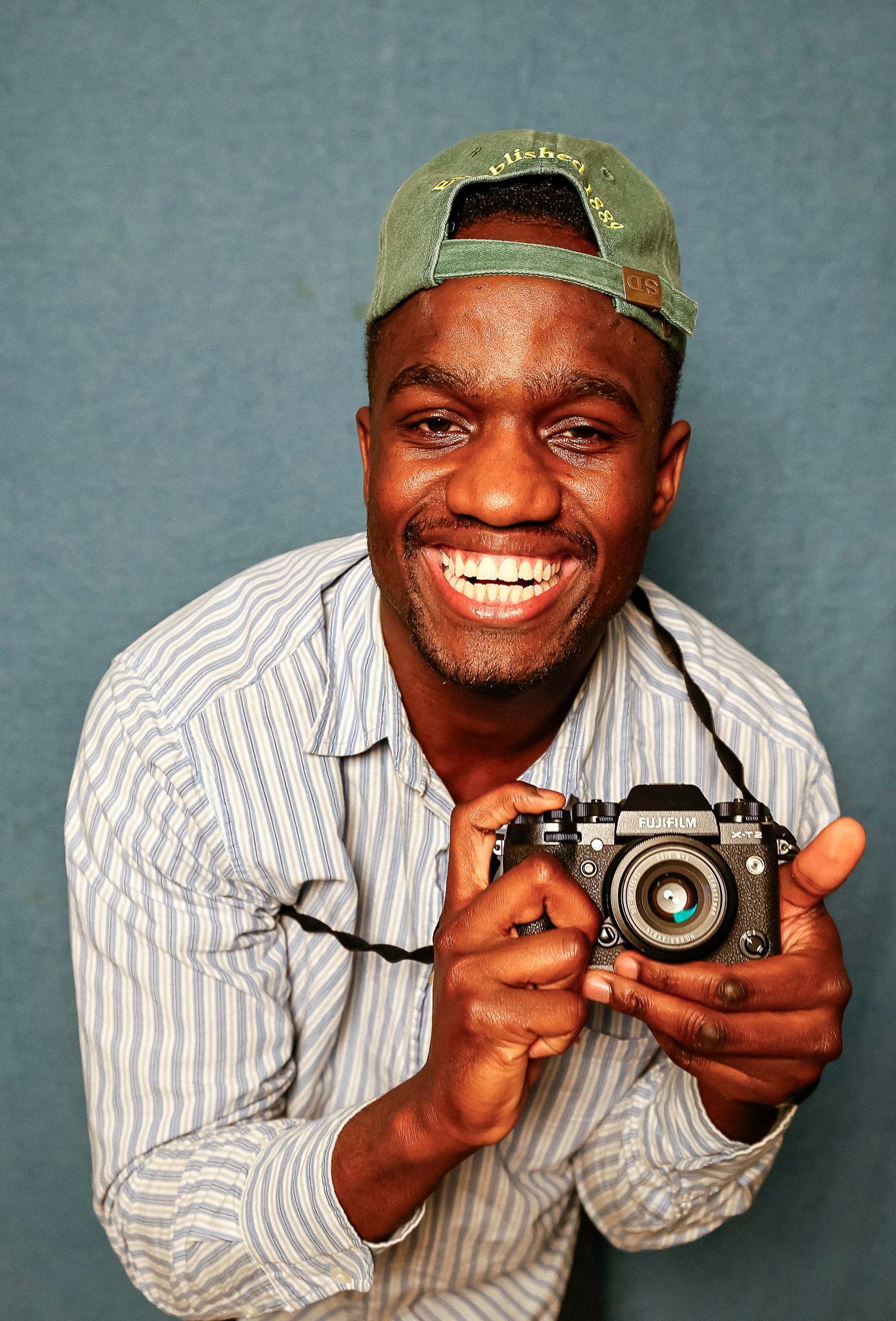 Smiling young man holding a film camera, wearing a striped shirt and green baseball cap against a blue background.