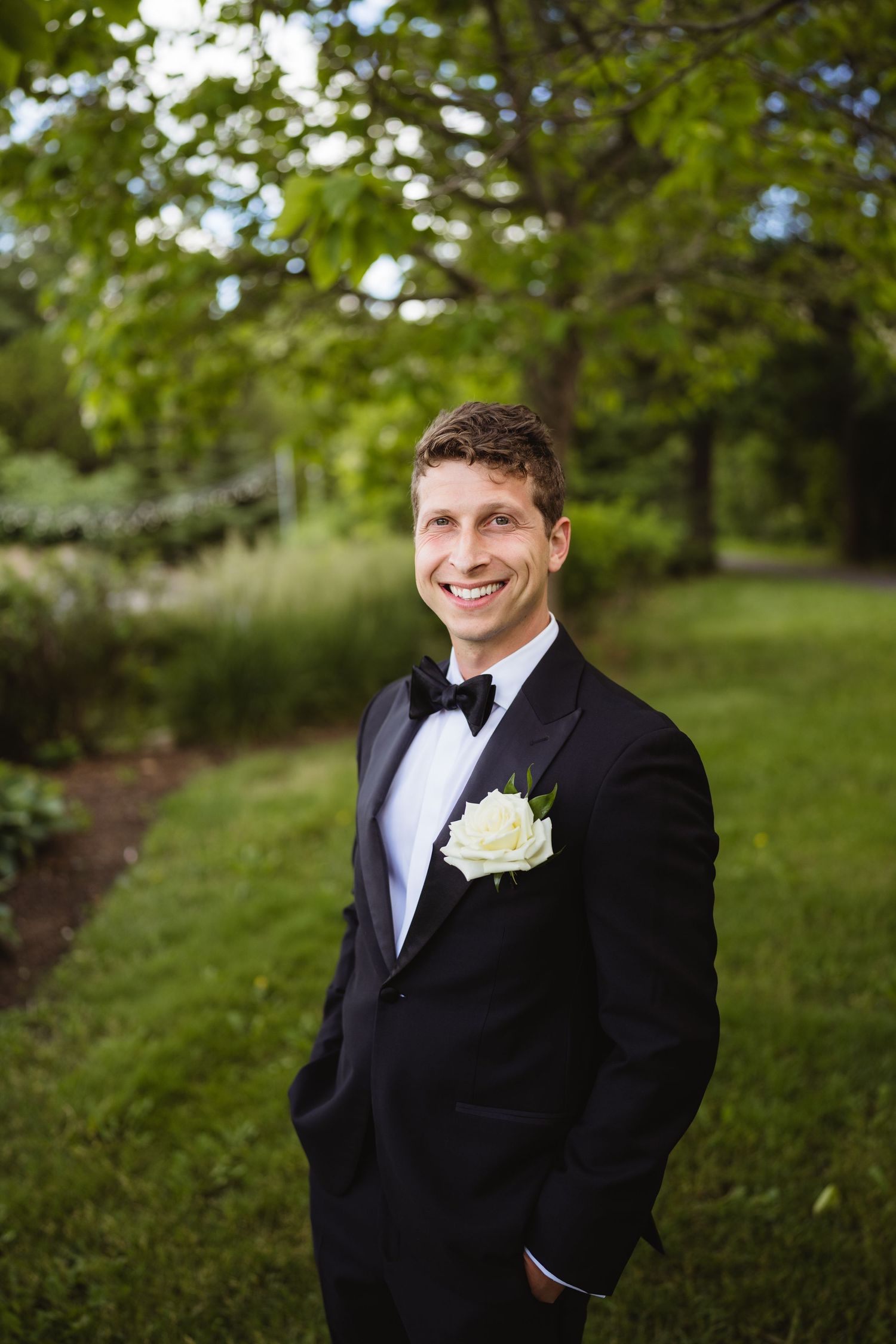 Smiling groom in black tuxedo with white rose boutonniere stands in lush green garden on wedding day.