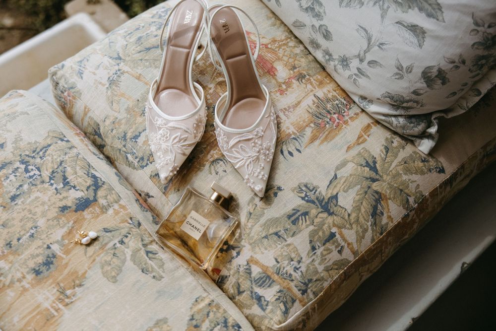 Delicate lace bridal heels displayed beside a perfume bottle on a floral patterned surface.