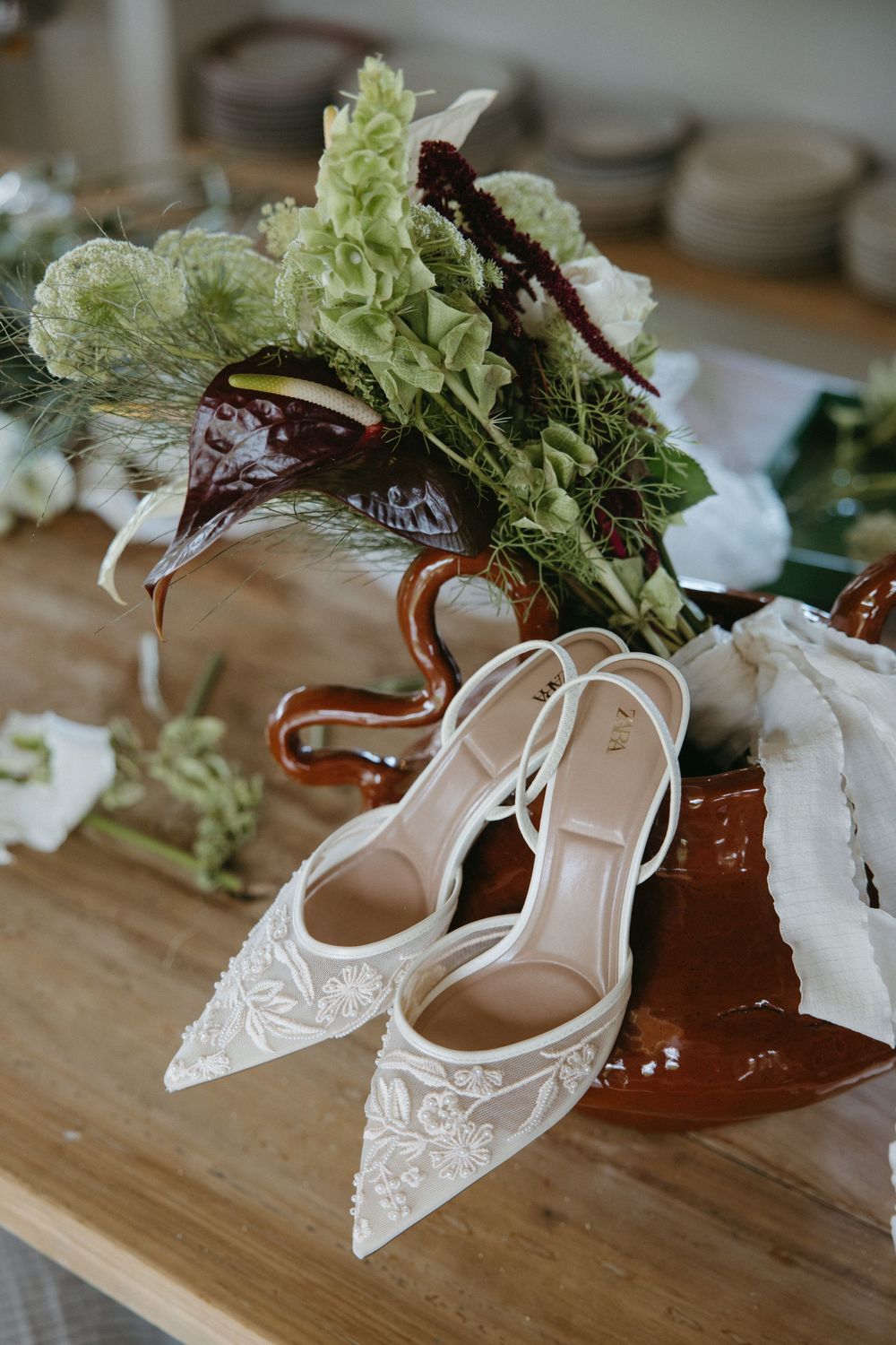 Elegant white lace bridal heels displayed on wooden table with rustic floral bouquet arrangement.