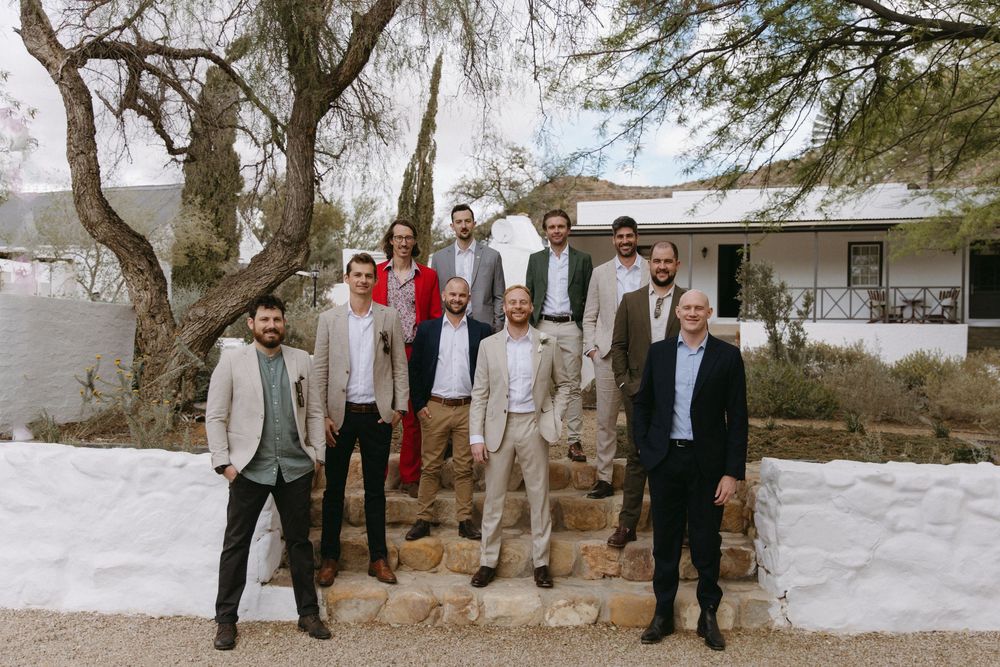 Group of wedding guests posing on stone steps outdoors near a rustic white building.