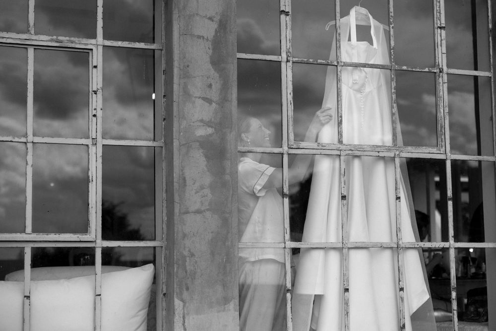 Black and white image of a wedding dress hanging by large industrial windows before the ceremony.
