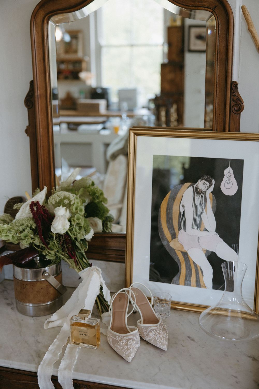 Elegant wedding flat lay with lace bridal shoes, bouquet, perfume, and framed art by vintage mirror.