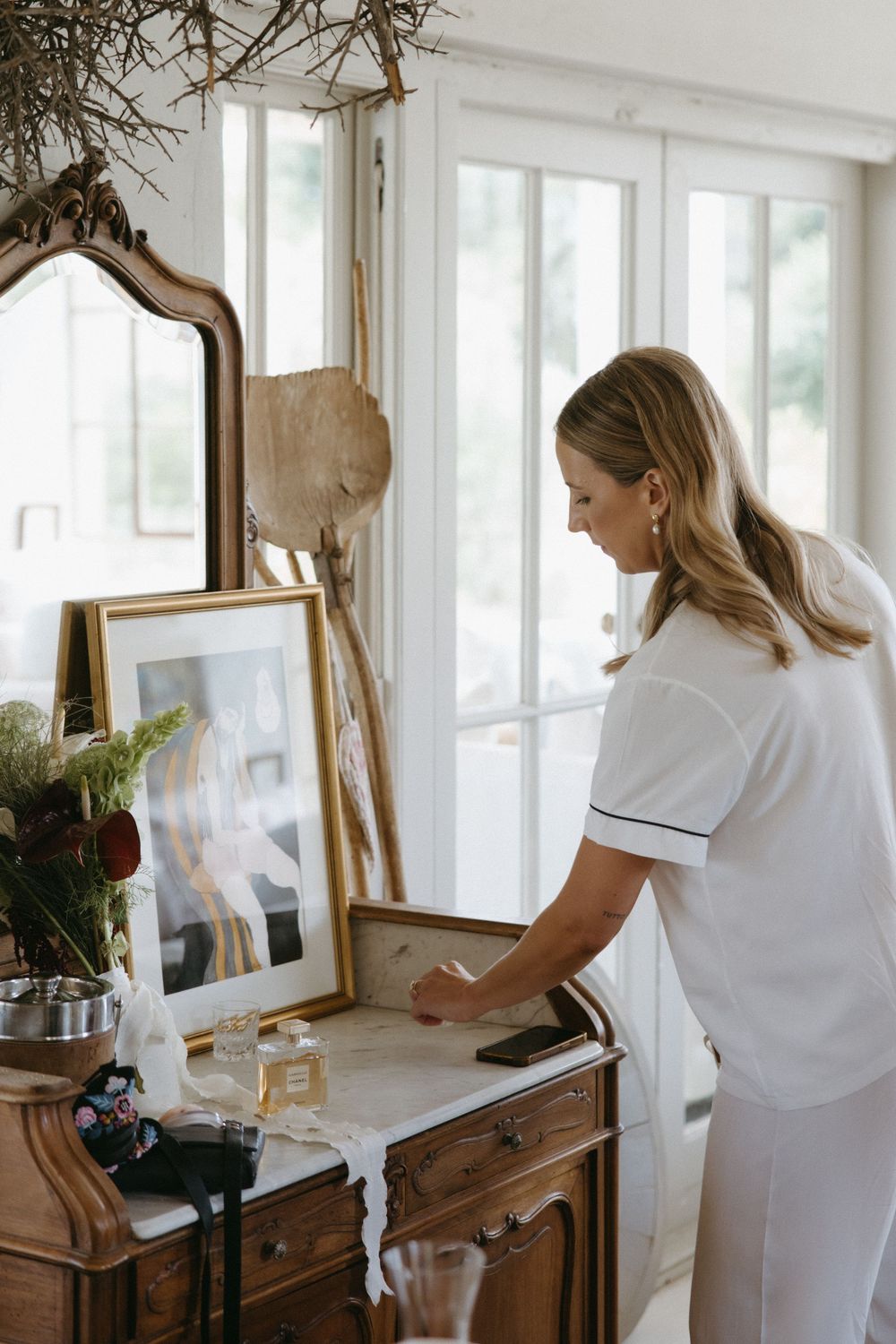Blonde bride in white robe preparing at a vintage wooden dresser with mirror before her wedding.