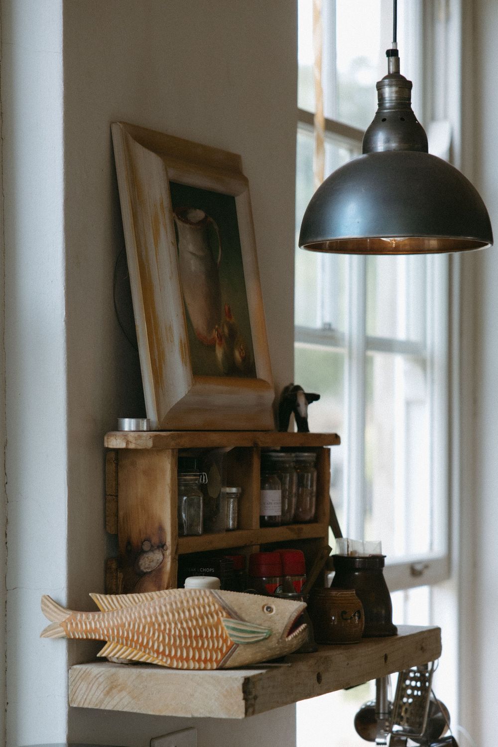 Rustic kitchen shelf with wooden spice cabinet, fish sculpture, framed artwork, and vintage pendant light near window.