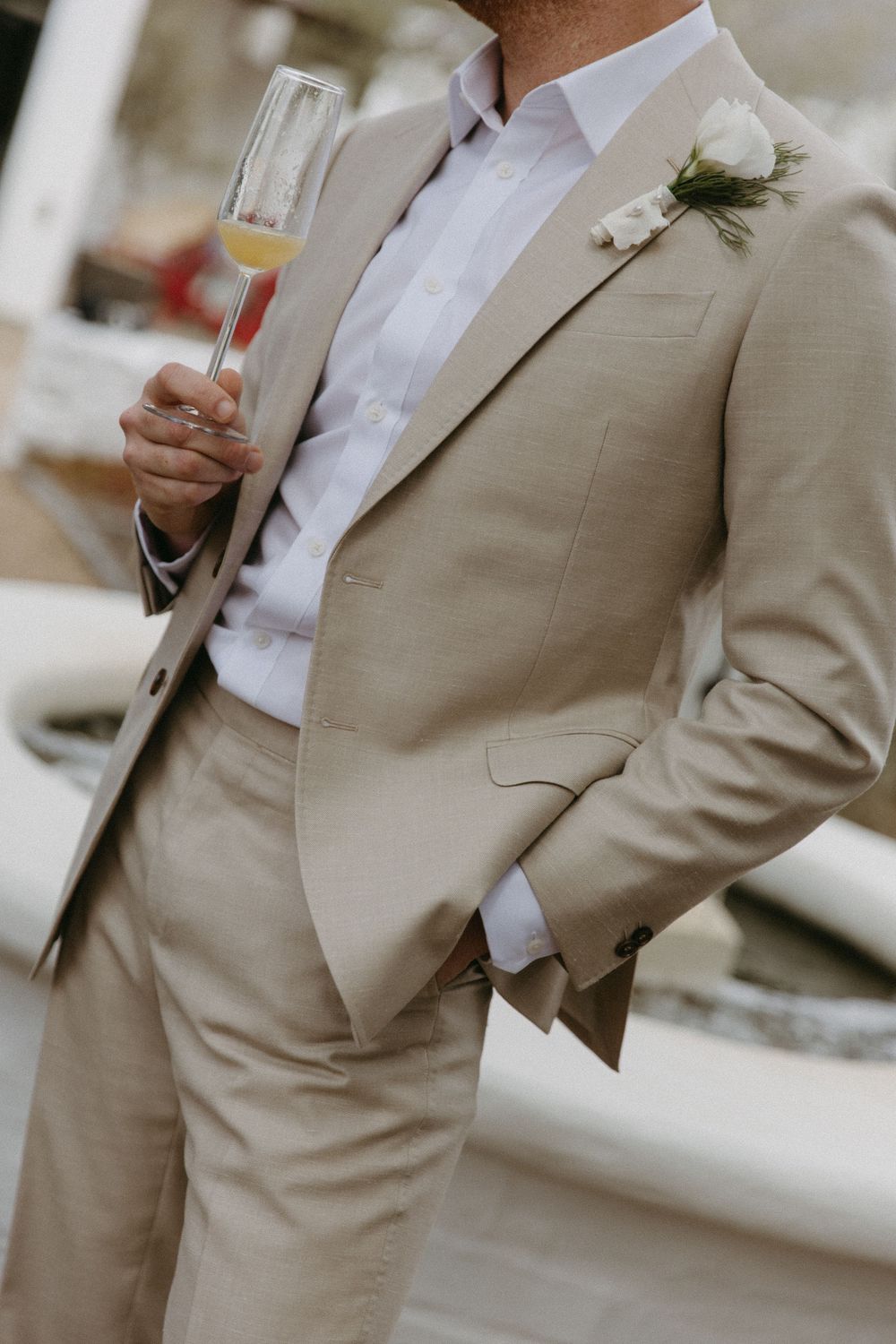 Groom in beige suit holding champagne flute with white boutonniere at wedding.