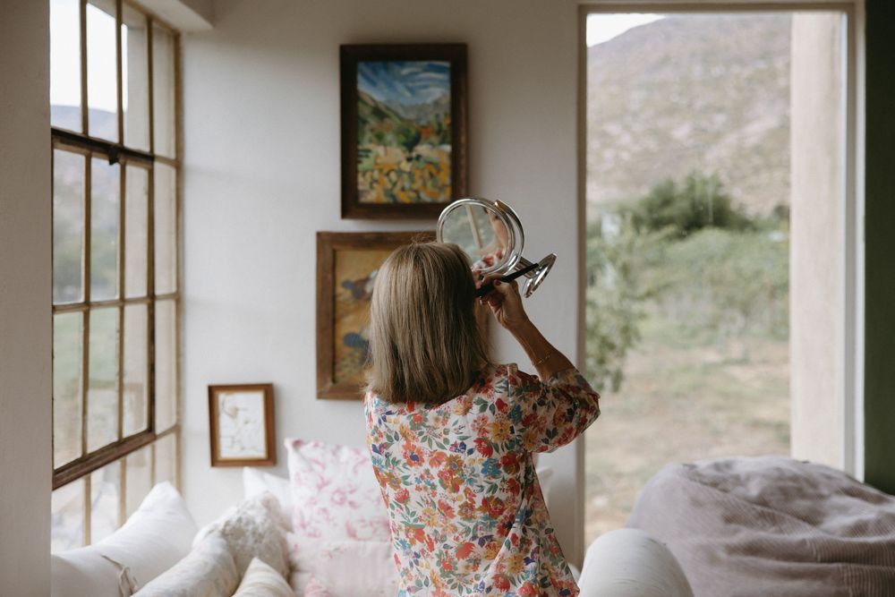 Woman in floral dress holding hand mirror near bright window in cozy room with landscape paintings.