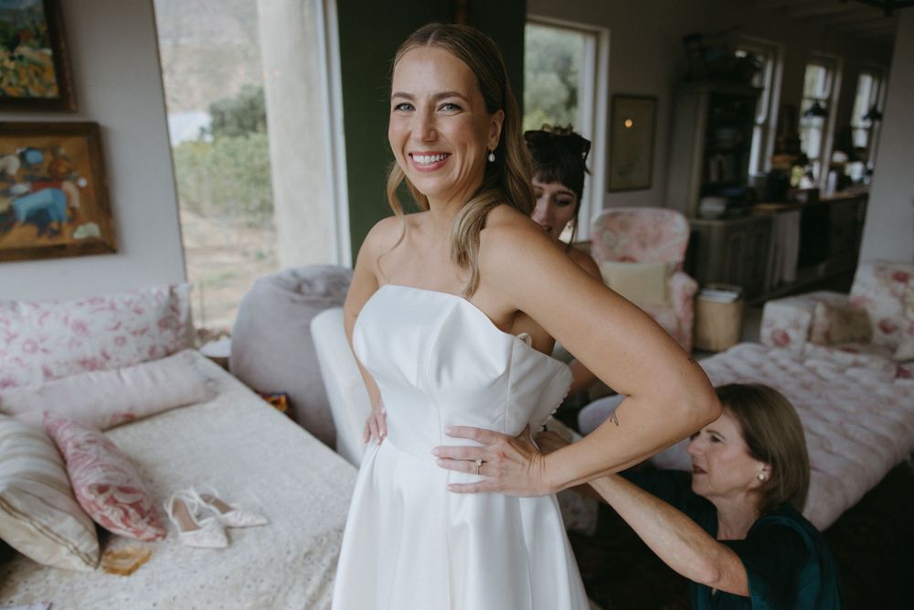 Smiling bride in strapless white gown being helped with dress by bridesmaid in cozy bedroom.