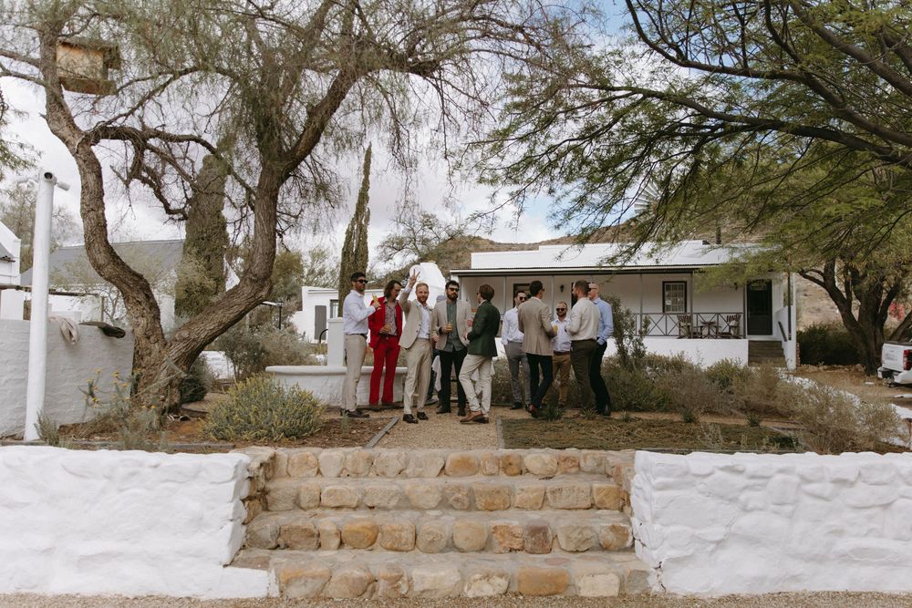 Wedding guests mingling outside a whitewashed farmhouse venue surrounded by desert trees and stone steps.